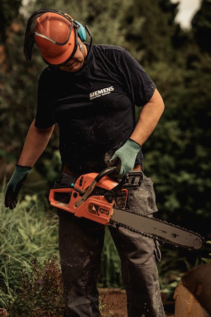A person engaged in outdoor garden maintenance, dressed in safety gear including a protective helmet, gloves, and a dark t-shirt with the Siemens logo, operating a chainsaw amidst a lush garden settin