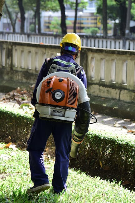 A person engaged in outdoor garden maintenance, dressed in safety gear including a protective helmet, gloves, and a dark t-shirt with the Siemens logo, operating a chainsaw amidst a lush garden settin