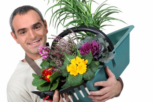 The image depicts a smiling man holding a blue plastic garden trowel and a black basket filled with a variety of colorful flowering plants, including yellow, purple, and red blooms, alongside lush green foliage. Behind him, part of a healthy green shrub or small tree is visible, suggesting a well-maintained outdoor garden space. The background features a clear, bright weather condition with natural light illuminating the scene. The garden setting appears neat and cultivated, with a focus on healthy plant growth suitable for outdoor landscaping and gardening services, such as hedge trimming and garden maintenance in Stratford or nearby areas. The man's attire is casual, appropriate for gardening work, and the image overall emphasizes the importance of healthy, vibrant plants and tidy outdoor environments, aligning with professional gardening offerings from companies like Hedge Trimming Stratford.